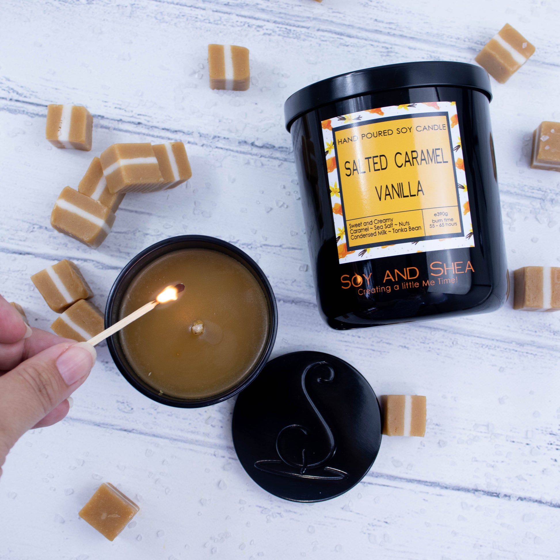 A flat lay photo showing a large black jar lying on its side on a white wooden surface surrounded by caramel sweets and sea salt.  To the bottom left of the jar is a smaller candle standing upright and filled with caramel coloured wax.  A hand reaches in with a lit match.