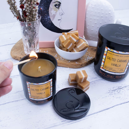 Two candles in black glass jars, with metal lids and decorative labels stand on a white wooded surface.   A larger looking candle sits to the right of the image with its lid on, and smaller one sits to the left with its lid removed and placed on the side revealing a caramel coloured wax.  A hand reaches in to light the candle with a match. In the background is a small white bowl of caramel pieces with sea salt scattered around, sitting next to a picture frame and vase of dried flowers.   