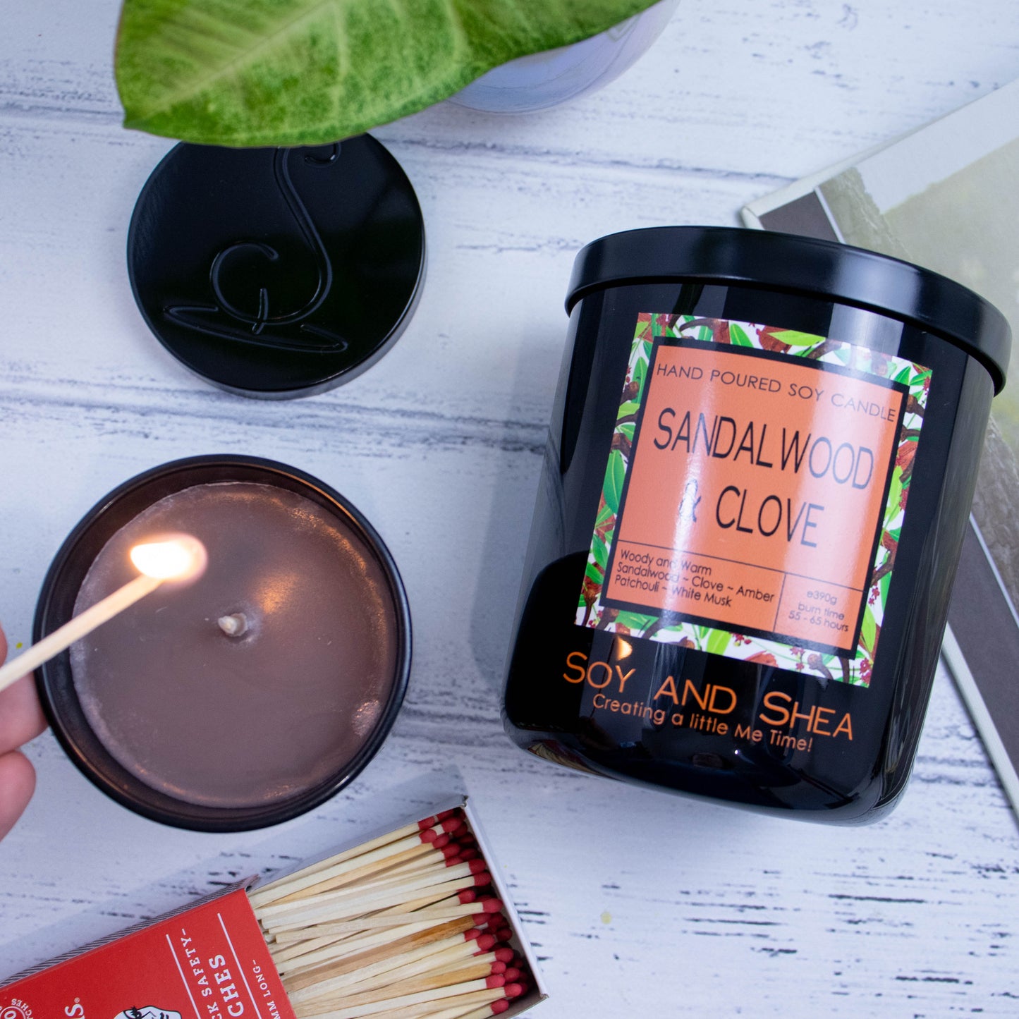 A flat lay photo showing a large black jar lying on its side on a white wooden surface next to a book and plant.  To the bottom of the jar is a smaller candle standing upright and filled with brown wax.  A hand reaches in with a lit match.