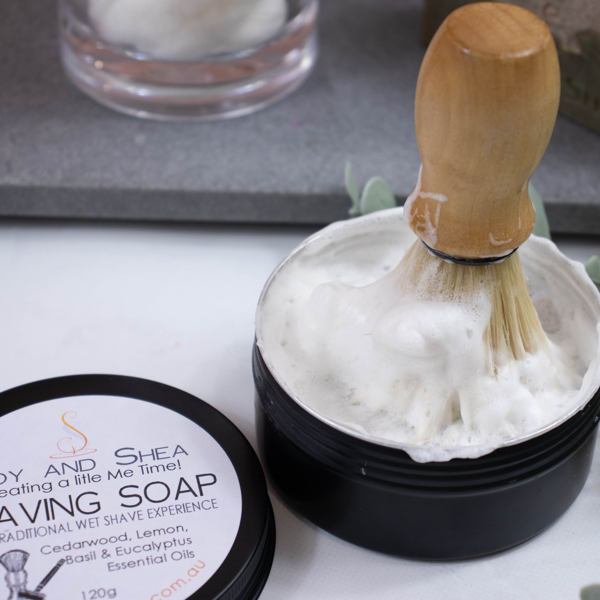 A pot of Shaving soap sits on the bench which has creamy lather. In the pot is a wooden handled shaving brush with foam over the bristles