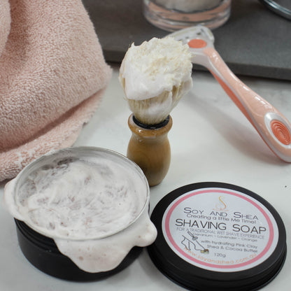 A black tin with its lid removed sits on a white bench with a dense lather over the top.  The lid lies next to the tin with a white round label with product details.  Behind the tin, a shaving brush with wooden handle stands upright on the bench and is covered in a dense lather.  In the back grouns is a pink towel and a razor propped on a stone tray.