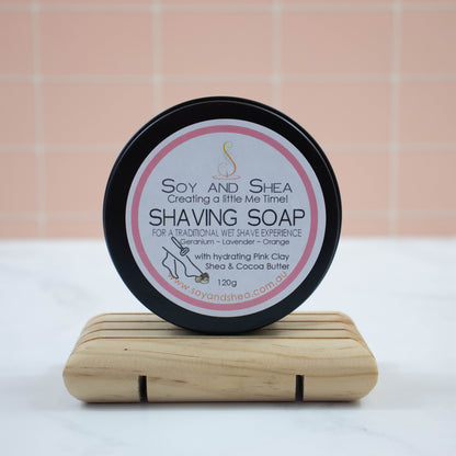 A black tin stands on its side on a wooden soap dish on a white background with peach tile background.  The lid of tin has a round white label with a pink border and text reading SHAVING SOAP FOR A TRADITIONAL WET SHAVE EXPERIENCE with a list of essential oils