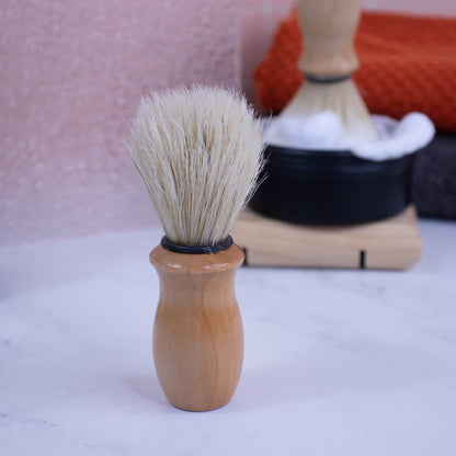 A shaving brush with wooden handle and cream coloured bristles stands upright on a white surface.  In the blurred background, a black tin with a dense foam can be seen with another brush standing in it with bristles in the foam.