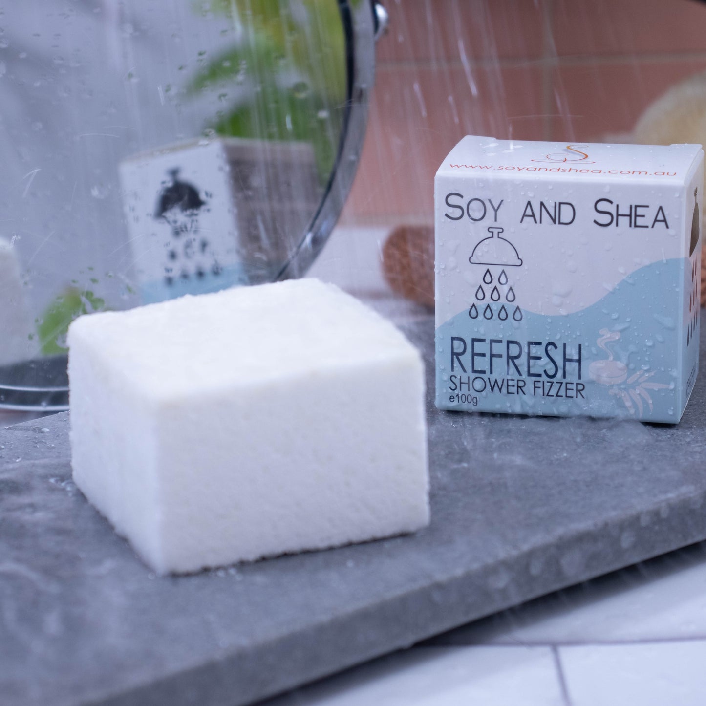 A shower of water falls over a white cube that is sitting on a grey stone slab. To the right of the cube is a white cardboard box, the same size, with a pale blue wave pattern . The box shows the details of the product. In the background, to the left, is a  mirror which reflects the box as well as  green plant.  To the right of the background is a pink tile wall and in front sits a wash cloth and brush.