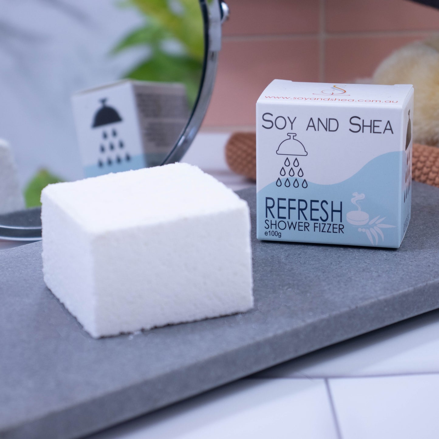 Sitting on a grey stone slab is a white cube and to its right is a white cardboard box, the same size, with a pale blue wave pattern . The box shows the details of the product. In the background, to the left, is a  mirror which reflects the box as well as  green plant.  To the right of the background is a pink tile wall and in front sits a wash cloth and brush.