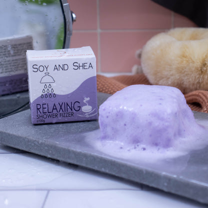A light purple cube sits on a stone slab and appears to be foaming. The product box sits to the left and has droplets of water on it as does the floor.  The box is white with a purple wave  design and shows details of the product.  In the background is a mirror reflecting the back of the box which has additional writing.  Hanging against the pink tile background is a grey towel and orange wash cloth with a dry body brush