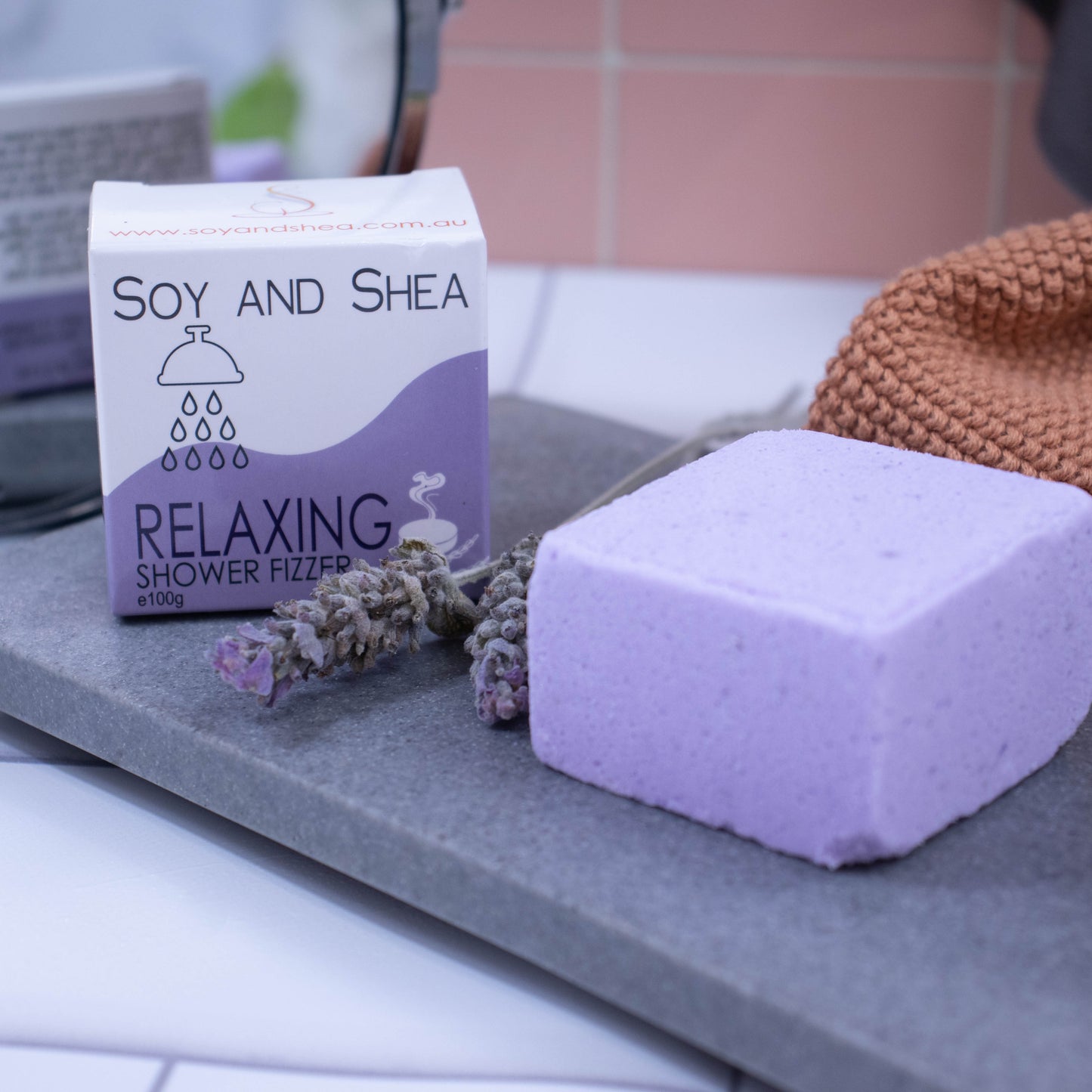 A light purple cube sits on a stone slab with a box of the product behind it.   Between them are two lavender flower spikes. The box is white with a purple wave design and shows details of the product. In the background is a mirror reflecting the back of the box which has additional writing.  Hanging against the pink tile background is a grey towel and a orange face cloth sits in front.