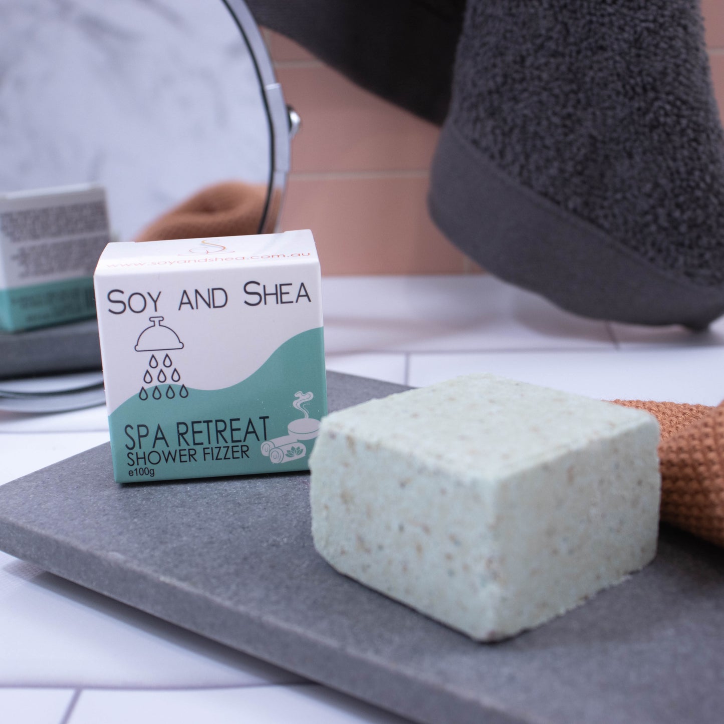 A light green cube with brown speckles sits on a stone slab with a box of the product behind it. The box is white with a sea green wave and shows details of the product. In the background is a mirror reflecting the back of the box which has additional writing.  Hanging against the pink tile background is a grey towel