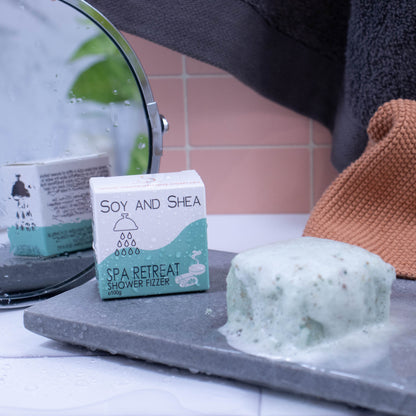 A light green cube with brown speckles sits on a stone slab and appears to be foaming. The product box sits to the left and has droplets of water on it as does the floor.  The box is white with a sea green wave and shows details of the product.  In the background is a mirror reflecting the back of the box which has additional writing.  Hanging against the pink tile background is a grey towel and orange wash cloth