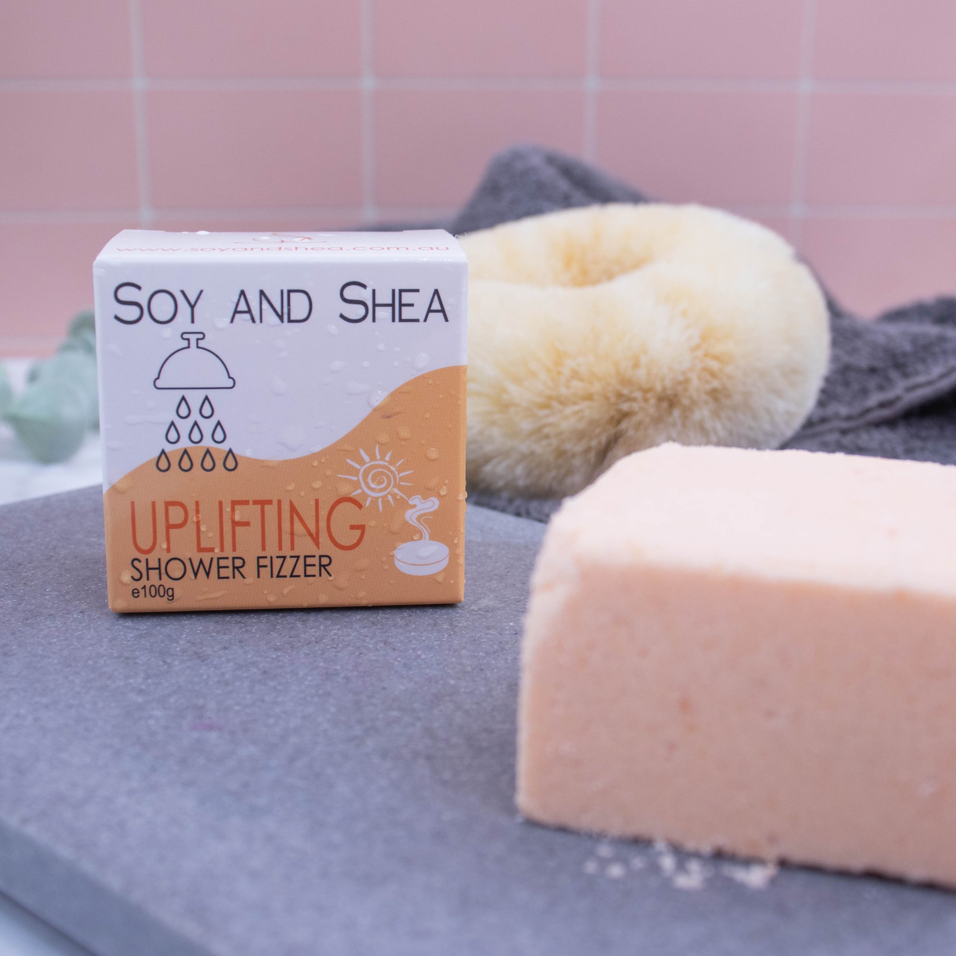 At the front of the picture sits a light orange block on a grey stone slab.  To the left and slightly back is the box which is white with a orange wavy pattern.  It has water droplets covering the box.  In the background is a pink tile wall along with a towel and body brush.