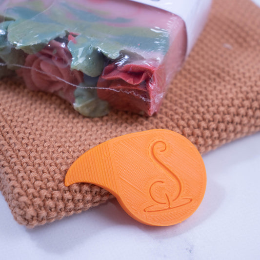 A bright orange shrinkwrap opener rests against the edge of a rust coloured face cloth upon which lies a red soap wrapped in plastic film.  The shrinkwrap opener is teardrop in shape with the top being pointed and curved.  It has the logomark engraved into it.