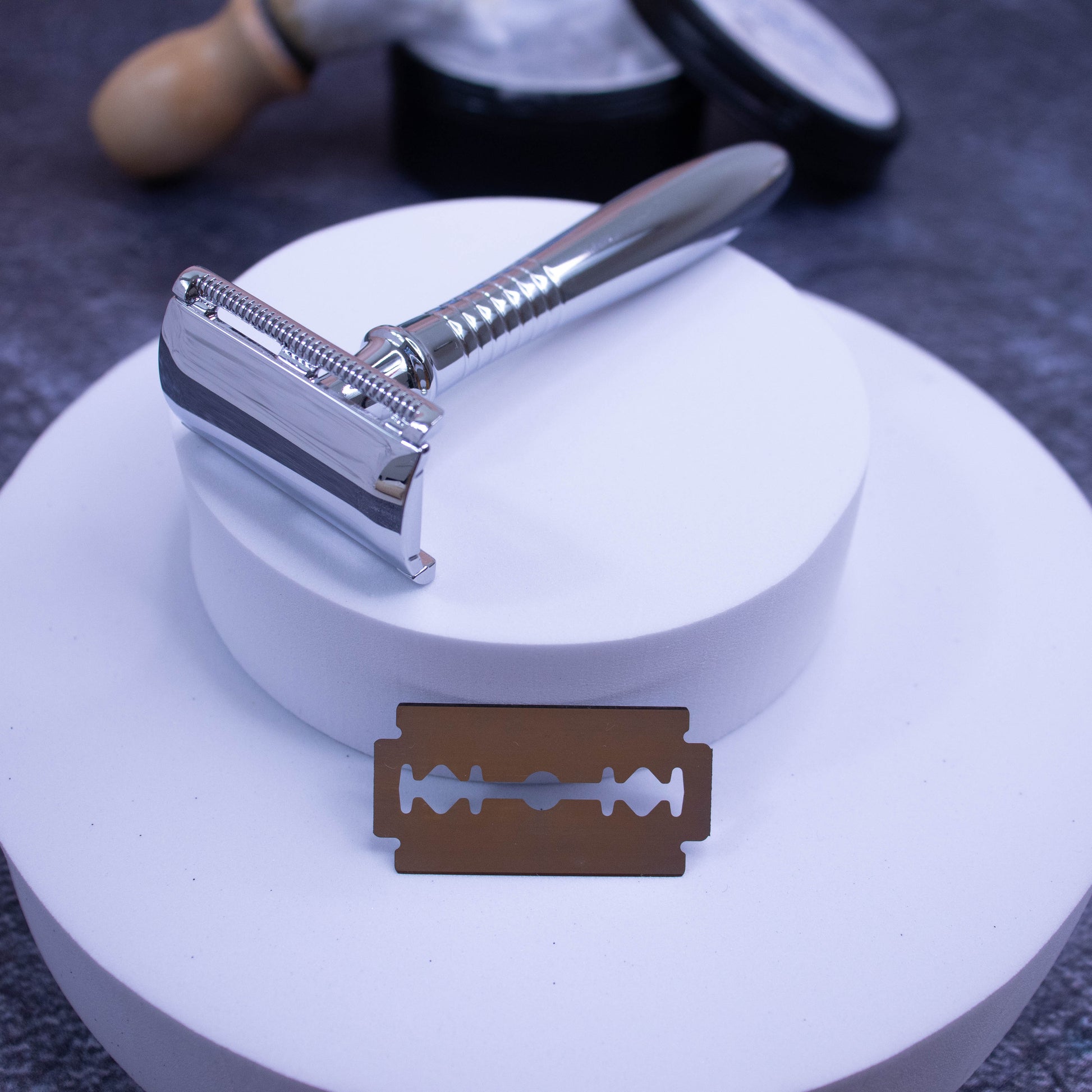 A bright silver safety razor rests on top of a stack of round foam pieces with a single razor blade resting against one layer below the razor.  The background is a dark surface with a blurred image of a tin of having soap and brush.