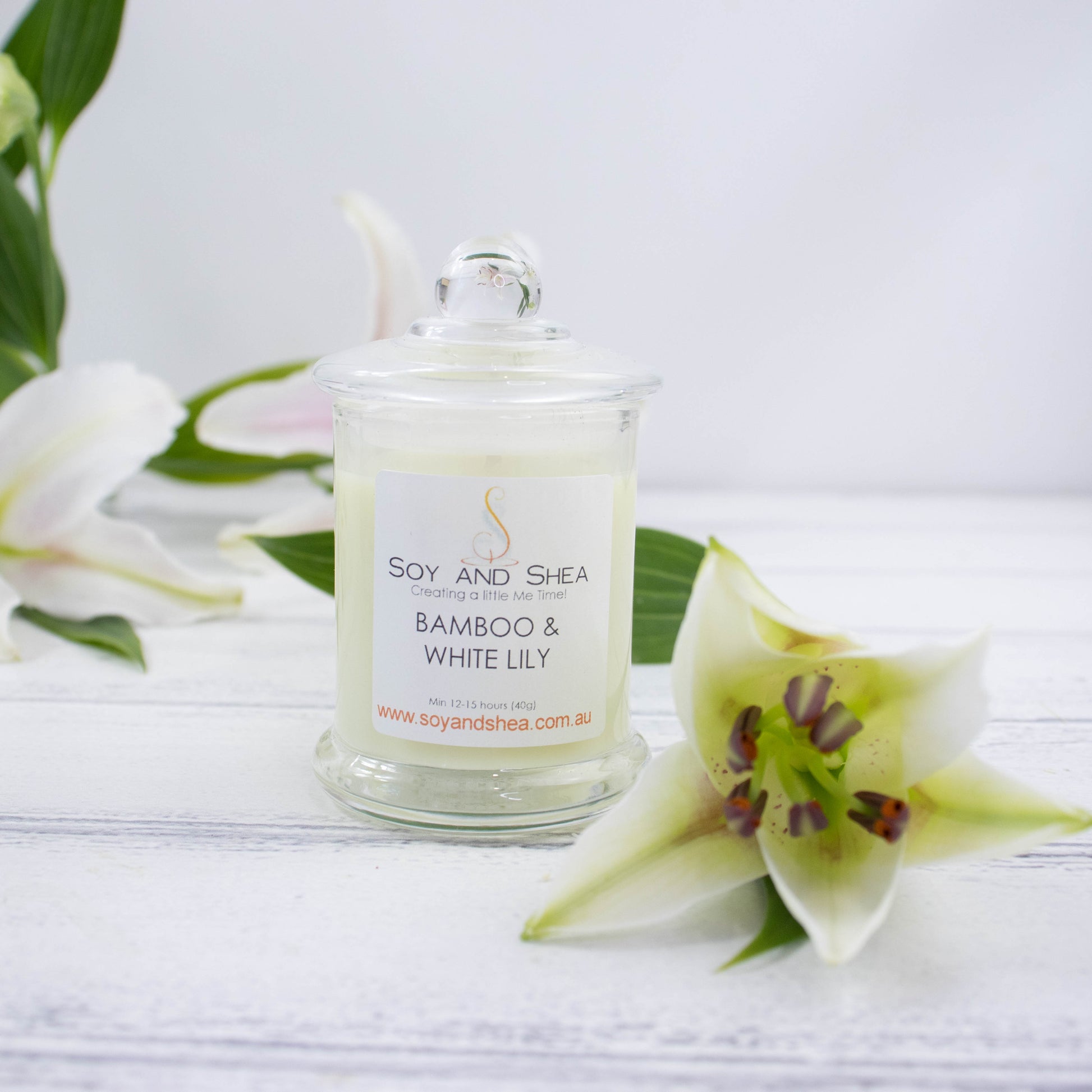 A small clear candle jar filled with light green wax stands on a white wooden surface featuring a glass lid finished with a ball.   It is surrounded by sprays of white lillies