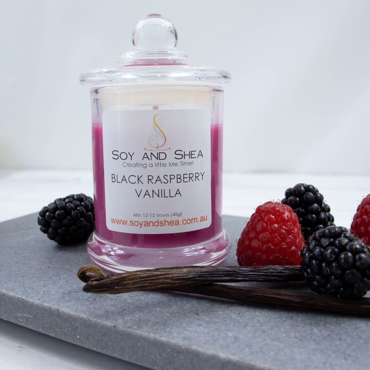 A small clear candle jar filled with magenta coloured wax stands on a grey stone  surface.   The jar has a glass lid with a ball shape top.  The candle is surrounded by black and red berries and two vanilla pods