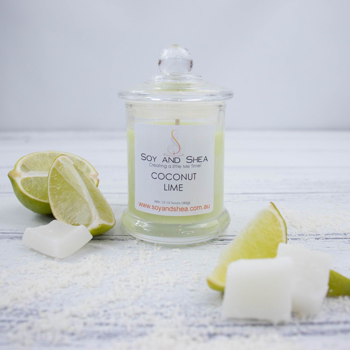 A small clear candle jar filled with light green wax stands on a white wooden surface surrounded by wedges of lime and pieces of coconut flesh.   The jar has a glass lid with a ball shape top.