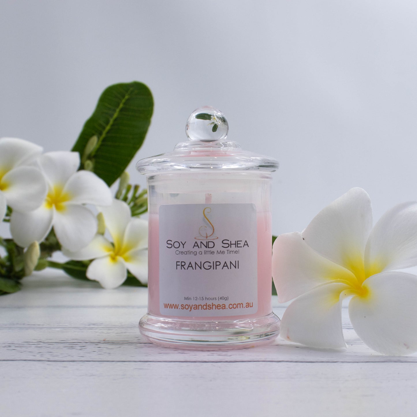 A small clear candle jar filled with liht pink wax stands on a white wooden surface.   The jar has a glass lid with a ball shape top.  The candle is surrounded by white frangani flowers with yellow centers and dark green leaves.