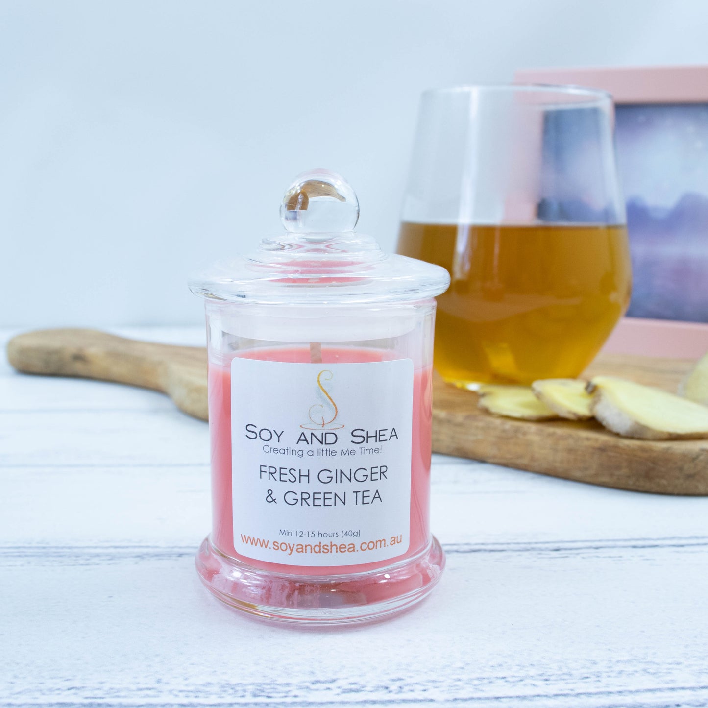 A small clear candle jar filled with red-orange wax stands on a white wooden surface.   The jar has a glass lid with a ball shape top.  In the background is a clear glass filled with tea and some slices of ginger