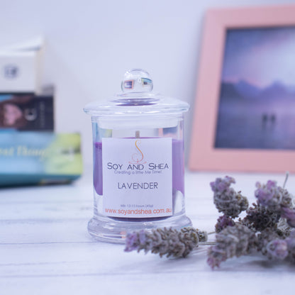 A small clear glass candle filled with purple wax sits on the wooden side surrounded by dry lavender.  The jar has a lid with a ball shape top. In the background is a picture and some books.