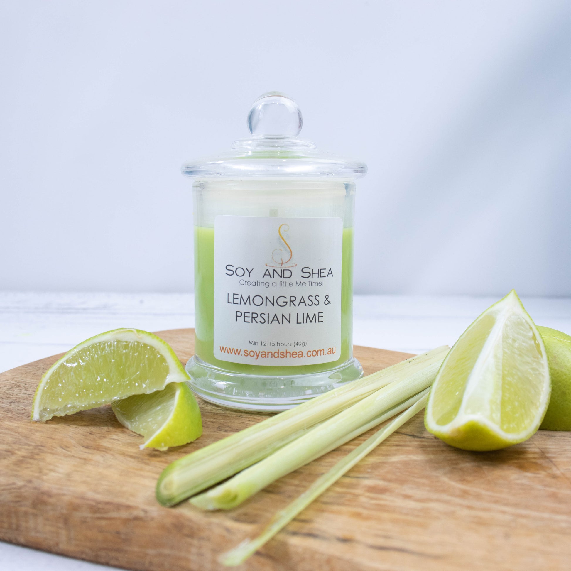 A small clear candle jar filled with bright green wax stands on a white wooden surface.   The jar has a glass lid with a ball shape top.   The candle is surrounded by wedges of lime and a piece of lemongrass.