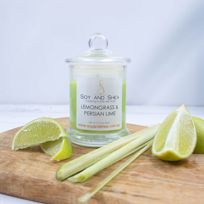 A small clear candle jar filled with bright green wax stands on a white wooden surface.   The jar has a glass lid with a ball shape top.   The candle is surrounded by wedges of lime and a piece of lemongrass.