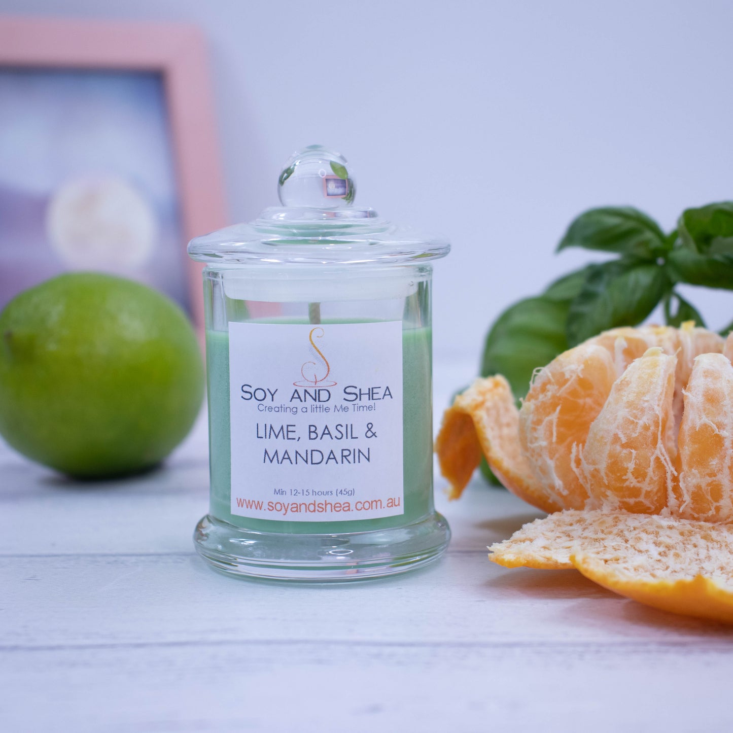 A small clear candle jar filled with green wax stands on a white wooden surface with a blurred background of a picture frame.  The jar has a lid with a ball shape top and is surrounded by a peeled mandarin, whole lime and bunch of basil.