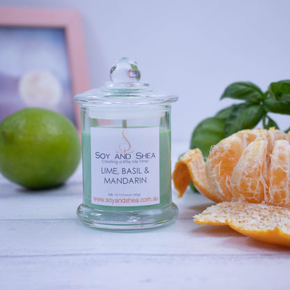 A small clear candle jar filled with green wax stands on a white wooden surface with a blurred background of a picture frame.  The jar has a lid with a ball shape top and is surrounded by a peeled mandarin, whole lime and bunch of basil.