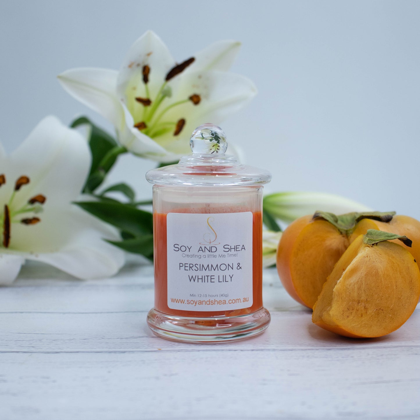 A small clear candle jar filled with deep orange coloured wax stands on a white wooden surface featuring a glass lid finished with a ball.  Next to the candle is a ripe persimmon with a wedge of fruit cut out and resting beside it.  Behind is a spray of white lily flowers.