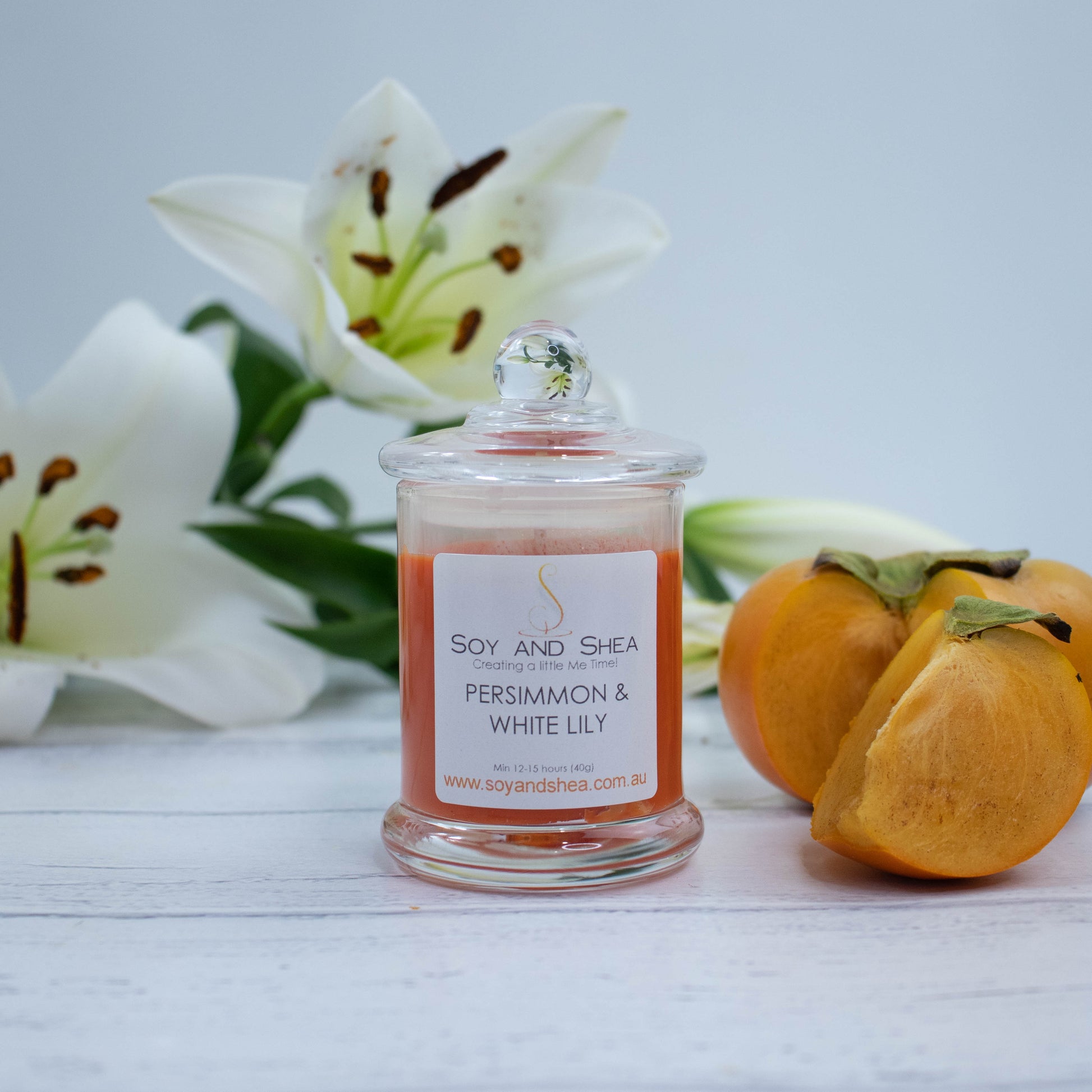 A small clear candle jar filled with deep orange coloured wax stands on a white wooden surface featuring a glass lid finished with a ball.  Next to the candle is a ripe persimmon with a wedge of fruit cut out and resting beside it.  Behind is a spray of white lily flowers.