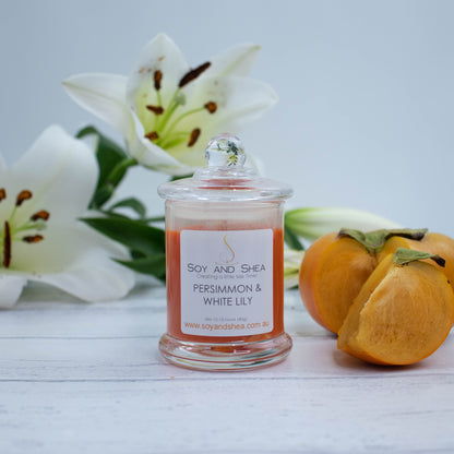A small clear candle jar filled with deep orange coloured wax stands on a white wooden surface featuring a glass lid finished with a ball.  Next to the candle is a ripe persimmon with a wedge of fruit cut out and resting beside it.  Behind is a spray of white lily flowers.