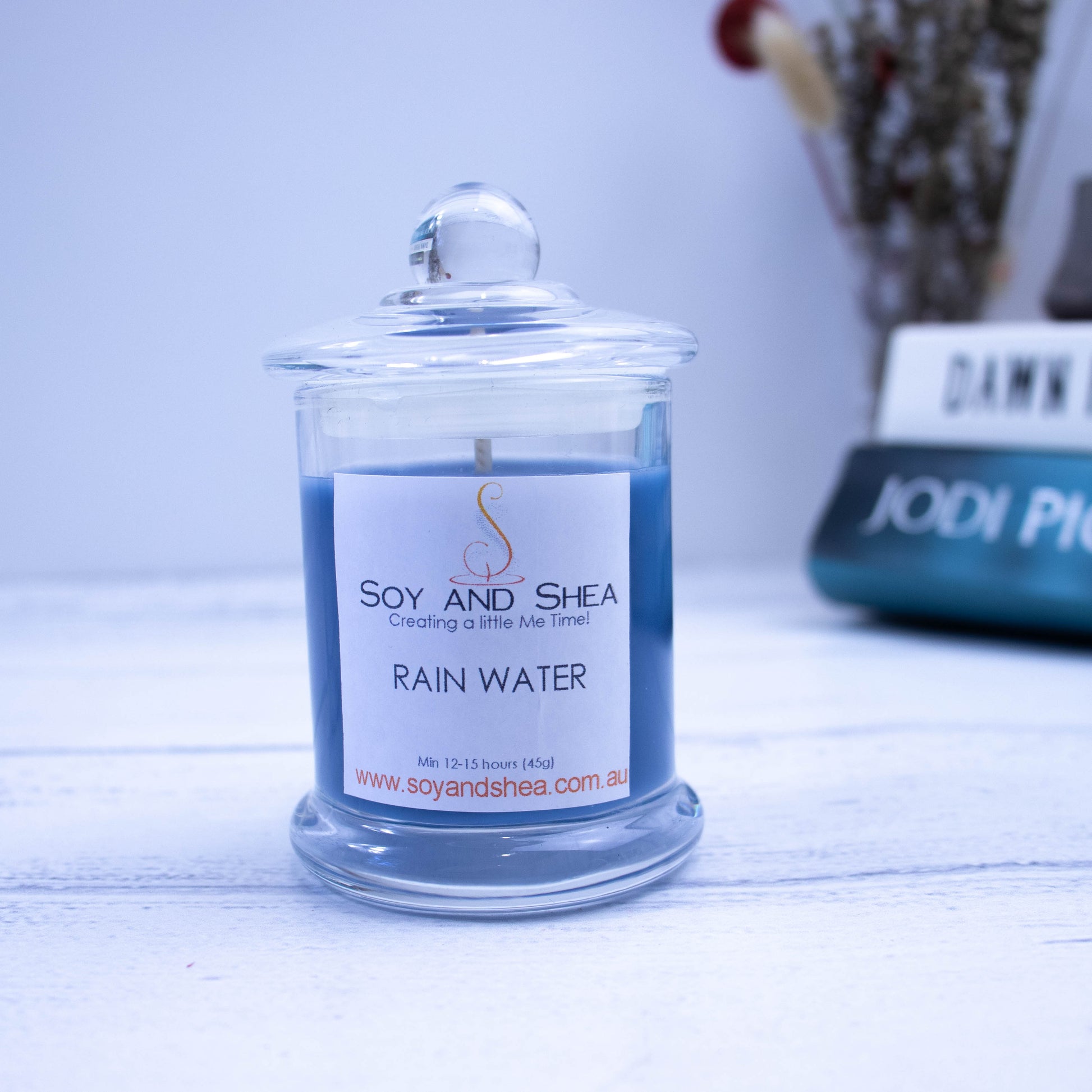 A small clear candle jar filled with dark blue wax stands on a white wooden surface with a blurred background of books, and vase with flowers.  The jar has a lid with a ball shape top.