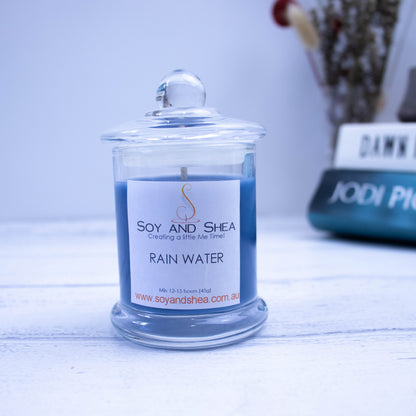 A small clear candle jar filled with dark blue wax stands on a white wooden surface with a blurred background of books, and vase with flowers.  The jar has a lid with a ball shape top.