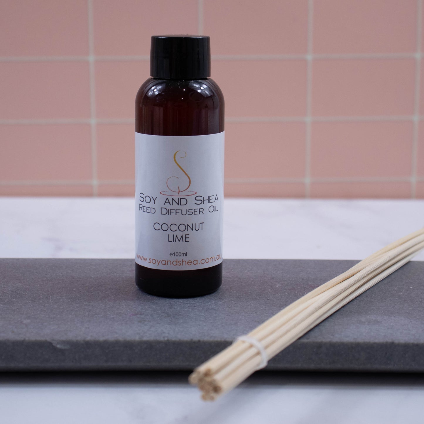 A brown bottle with black cap stands on a grey stone tray with a bundle of natural coloured reeds against a peach tile background.  It has a white label with text REED DIFFUSER OIL 100ml
