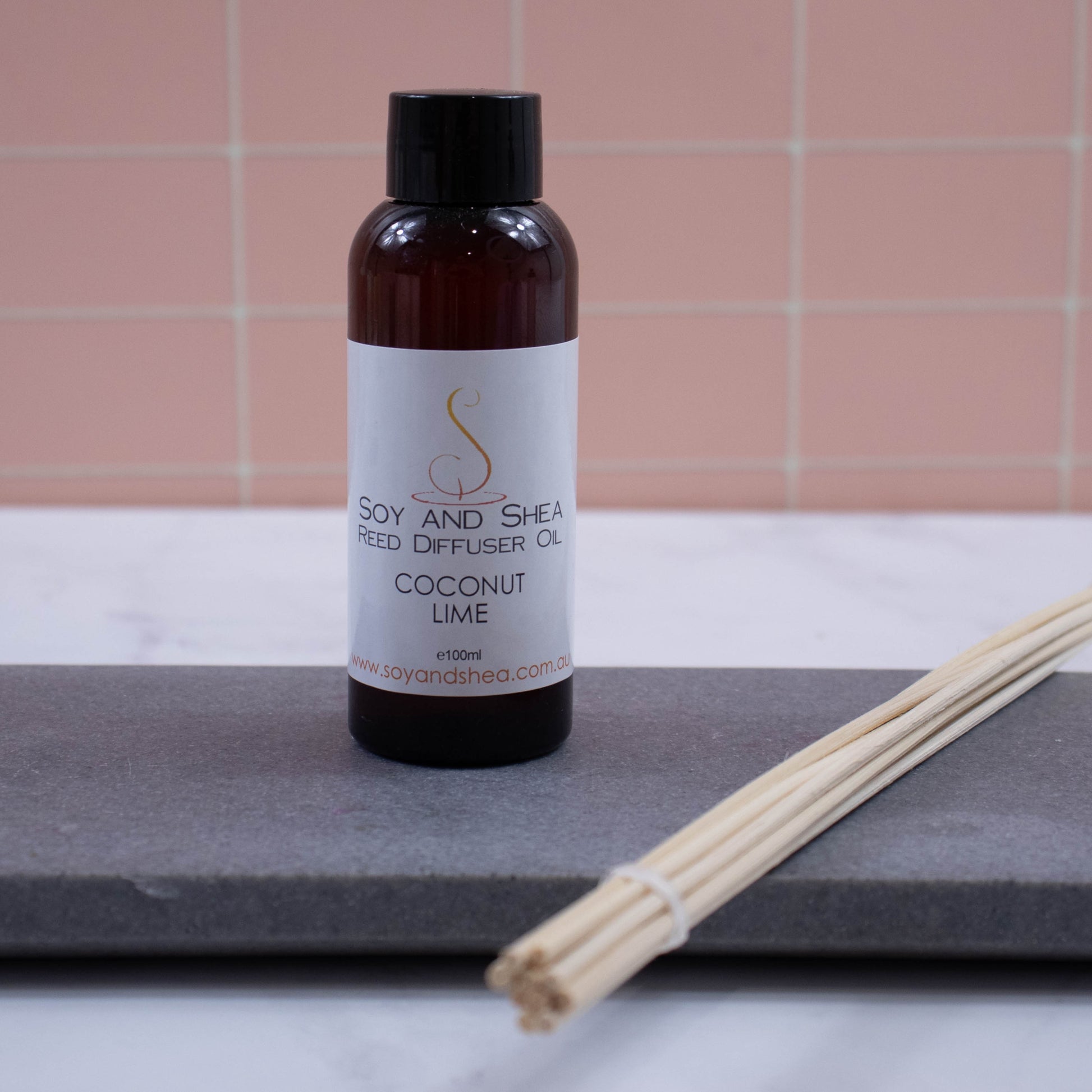 A brown bottle with black cap stands on a grey stone tray with a bundle of natural coloured reeds against a peach tile background.  It has a white label with text REED DIFFUSER OIL 100ml