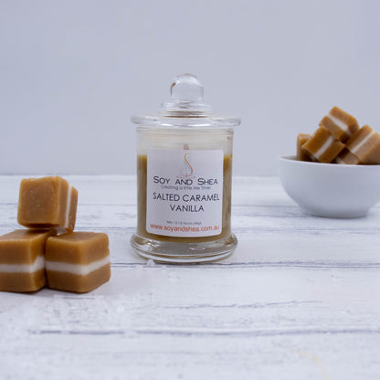A small clear candle jar filled with caramel coloured wax stands on a white wooden surface featuring a glass lid finished with a ball.    To the left is a pile of three caramels each with a white centre.  In the backgroudn is a small white bowl filled with more caramels.