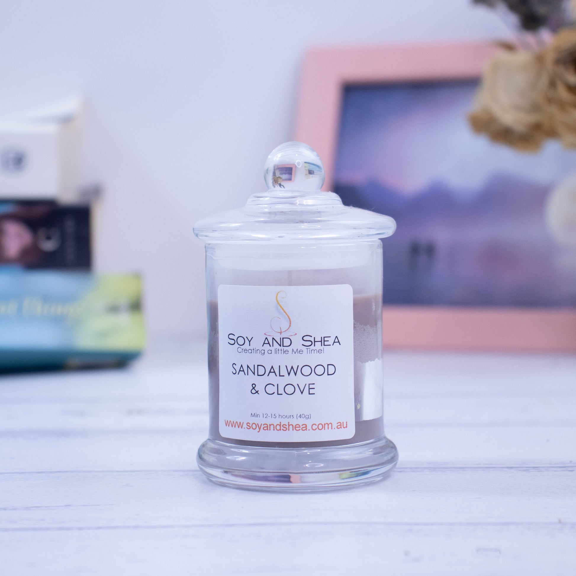 A small clear glass candle filled with light brown wax sits on the wooden side.  The jar has a lid with a ball shape top. In the background is a picture and some books.