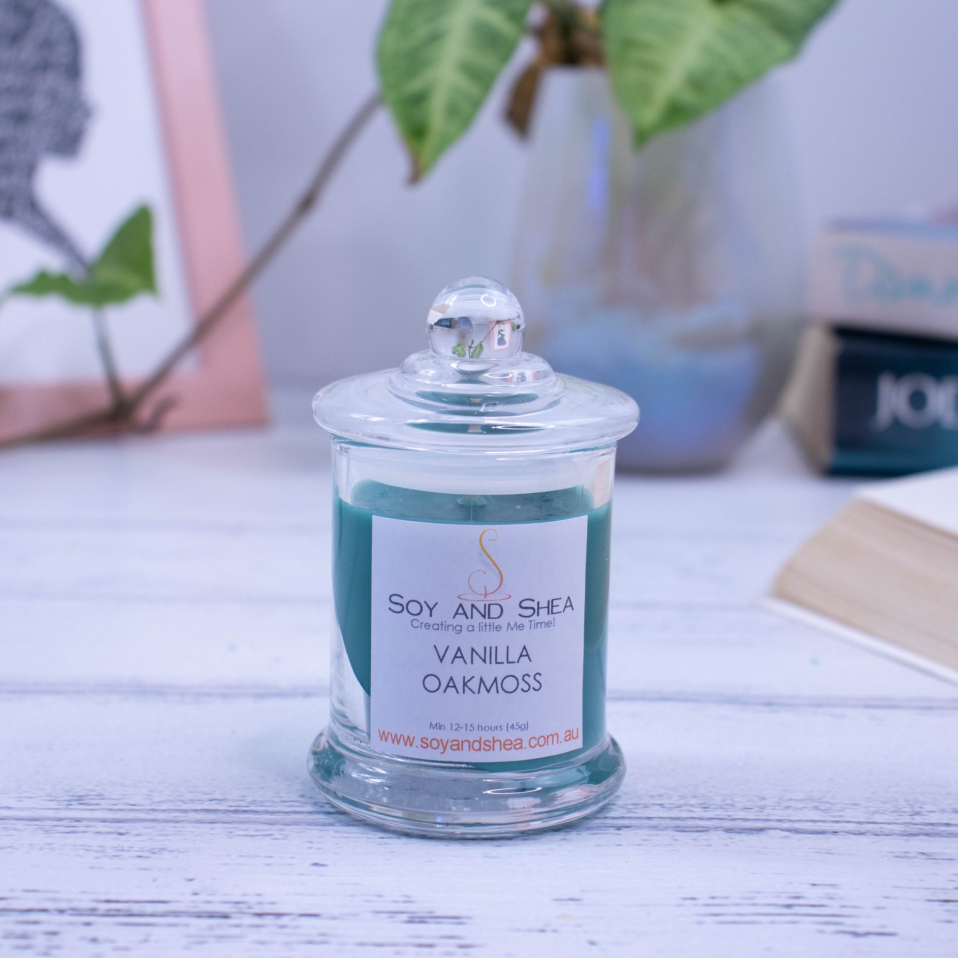 A small clear candle jar filled with dark green wax stands on a white wooden surface with a blurred background of books, picture frame and vase.   The jar has a lid with a ball shape top.
