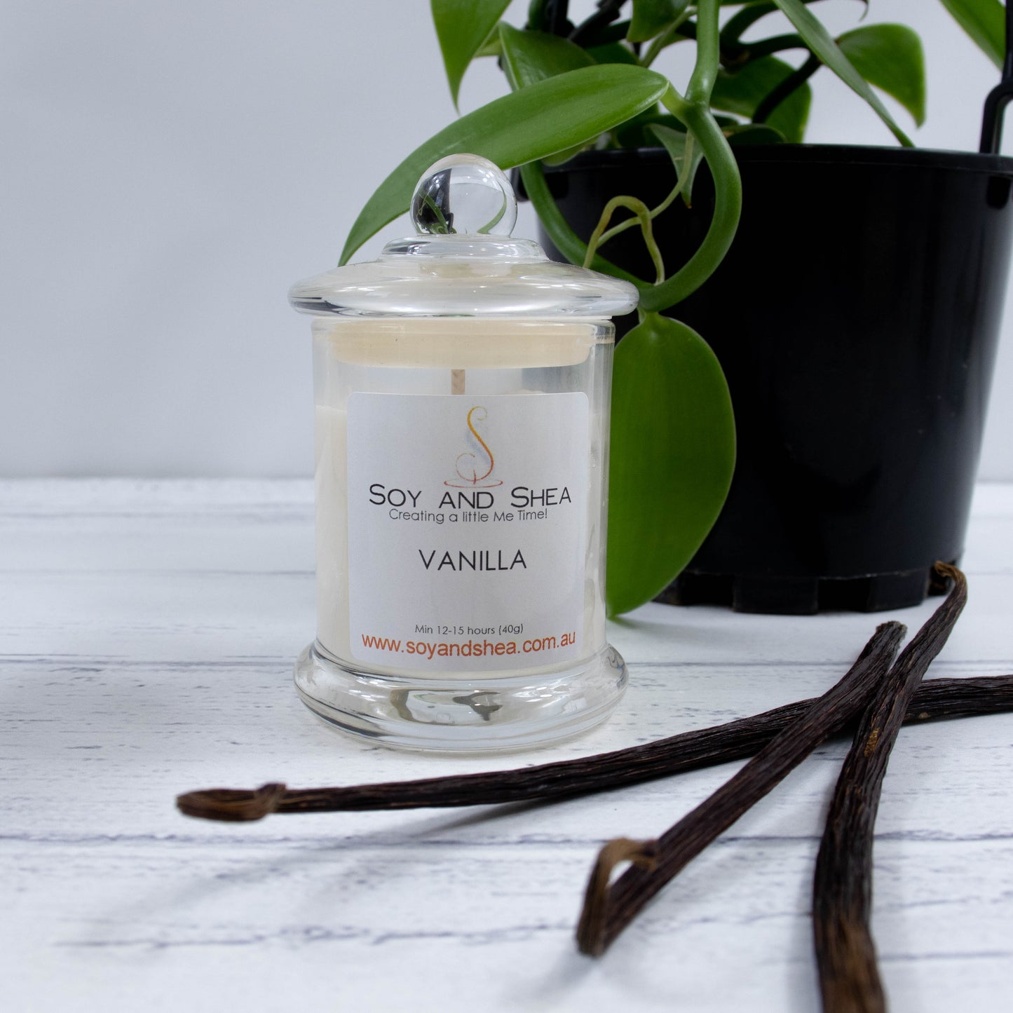 A small clear candle jar filled with reddish wax stands on a white wooden surface.   The jar has a glass lid with a ball shape top.  Behind the candle is a vanilla vine in a black pot and three vanilla pods lie beside the candle.