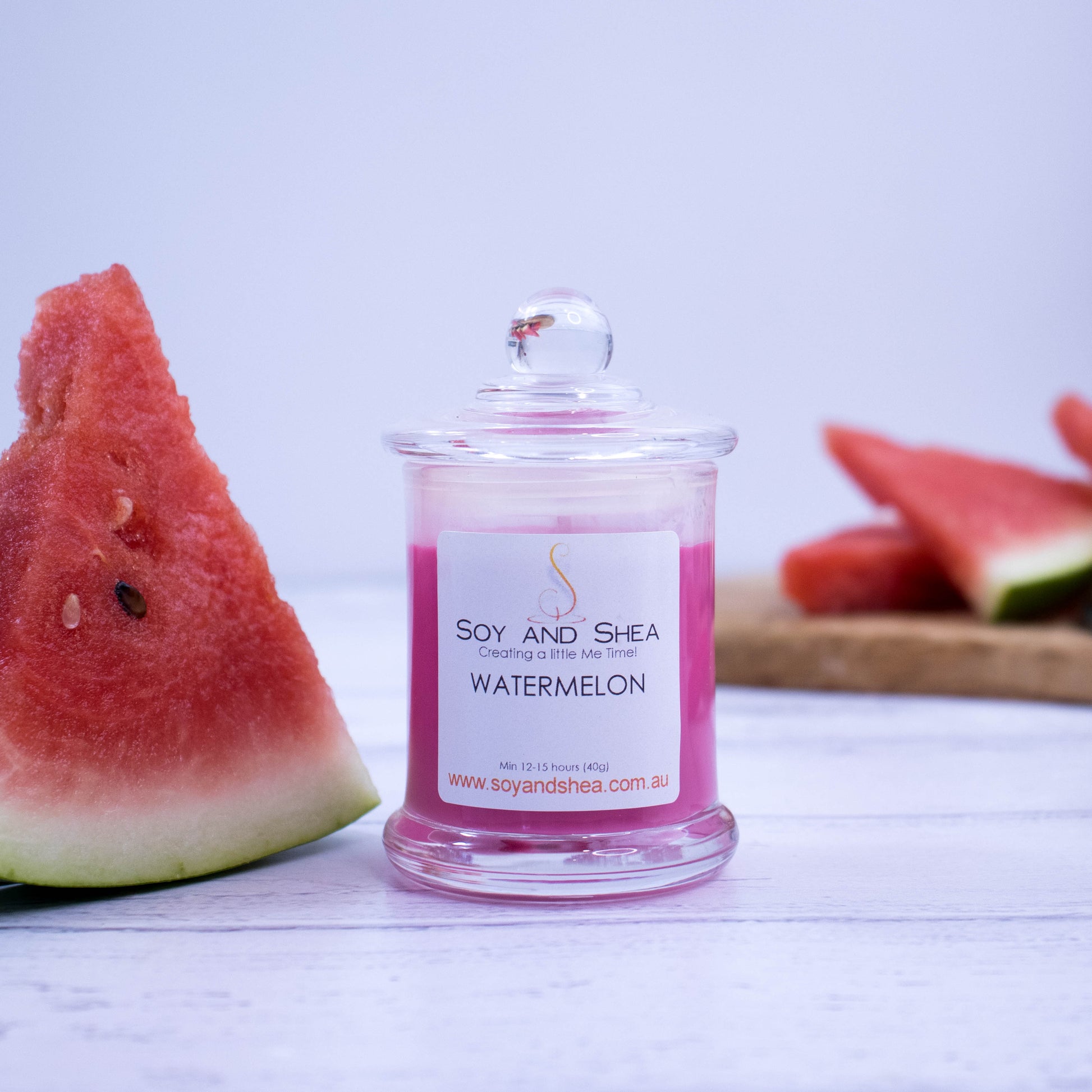 A small clear candle jar filled with magenta coloured wax stands on a white wooden surface next to a slice of watermelon.  More watermelon are resting on a wooden board in the background.
