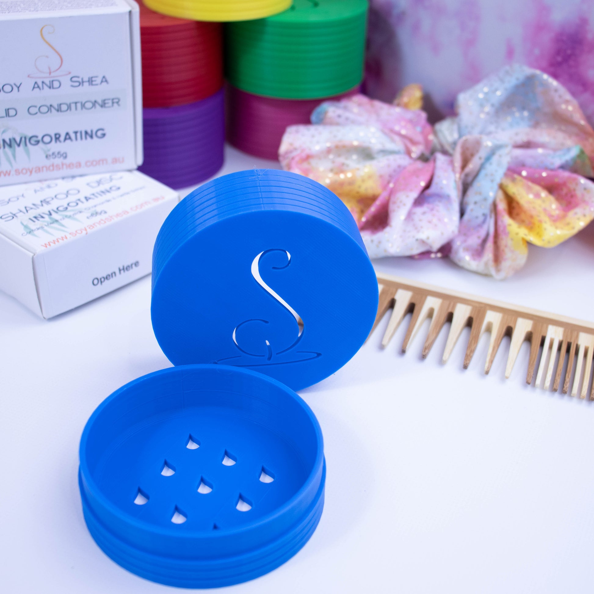 A bright blue shampoo pot base sits on a white surface with the lid standing behind on its sit.  The base shows cutout shapes of waterdrops while the top has the logomark cutout of it.  Behind is some Shampoo box packaging along with a scrunchie, wooden comb and stack of colourful Shampoo containers.