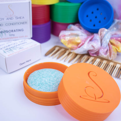 In the foreground sits an orange shampoo travel pot, a cylindrical container that splits into two.  The base of the pot holds a solid shampoo while the lid rests against the side with a cutout of the logomark.  In the background, a stack of colourful pots can be seen along with a wooden comb, scrunchie and shampoo packaging.