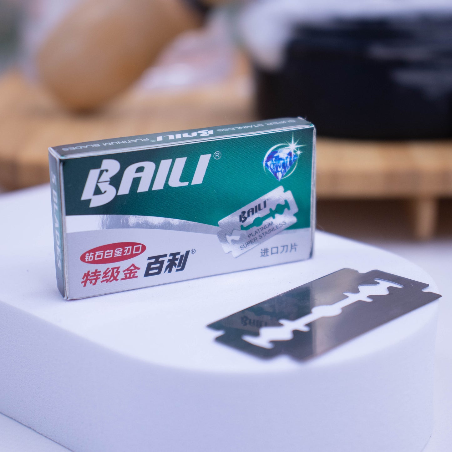A small box of Stainless Steel razor blades stands upright on a white surface with a single blade lying in front against a blurred background.