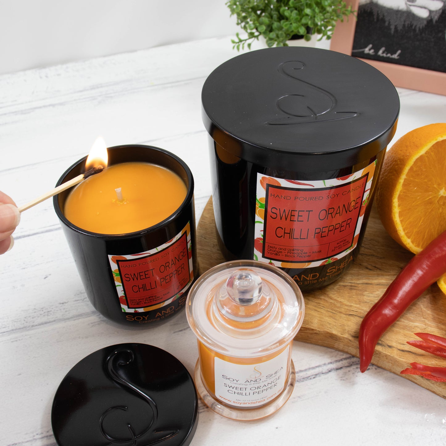 A collection of three different size candles are displayed on a wooden bench along with an orange that has been cut in half and a red chilli on a wooden board.  Next to the food is the largest of the candles which is a black glass jar with a metal lid showing the debossed logomark.  To the left is a small version with the lid removed to show an orange wax.  A hand reaches in to light it with a match.  In front is the smalles candle with is clear glass with decorative clear ball on the lid