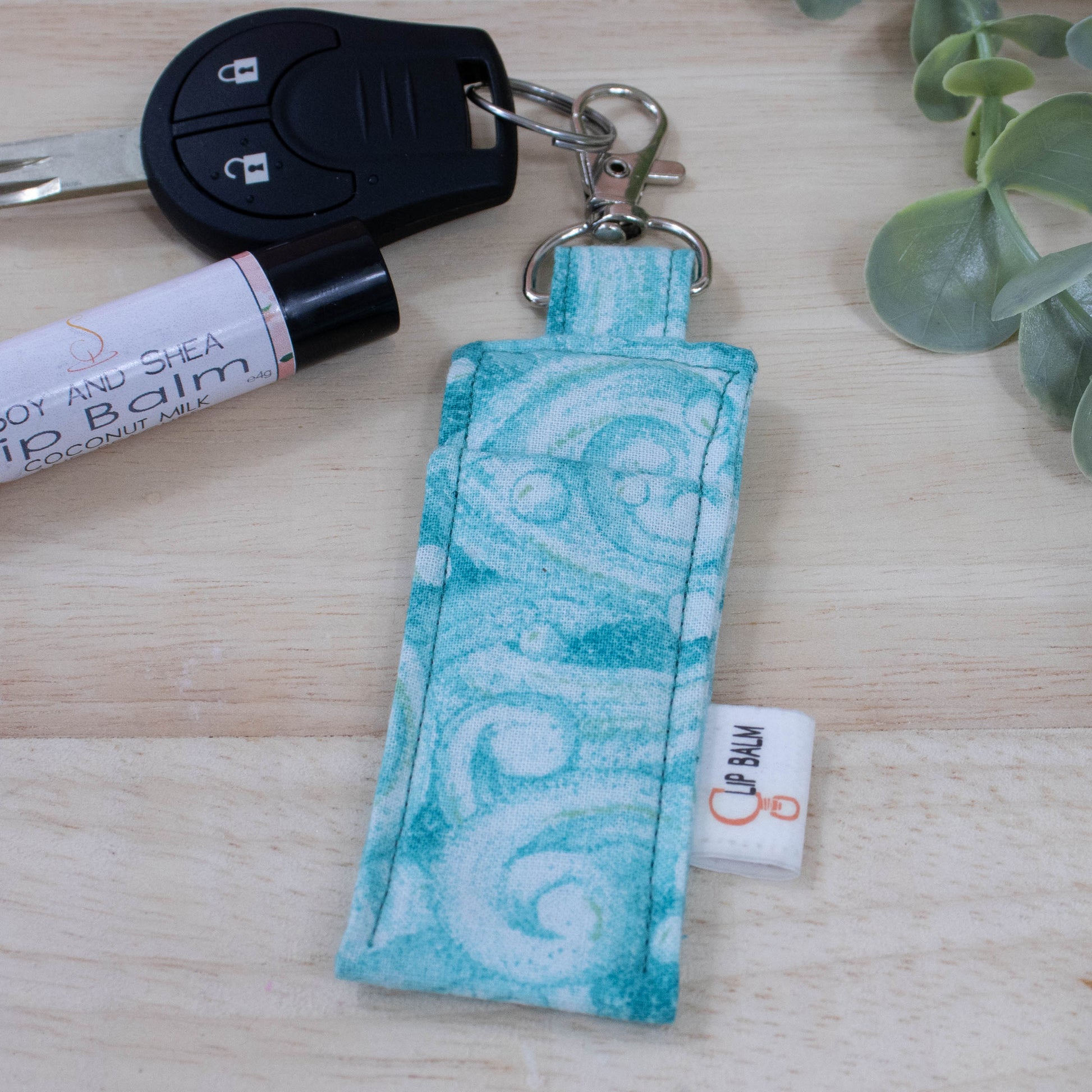 A clip balm holder lies on a wooden surface attached to a car key with a "lobster claw" clip.  To the side is a tube of lip balm and some greenery.  The Clip Balm has a watery wave pattern in teal
