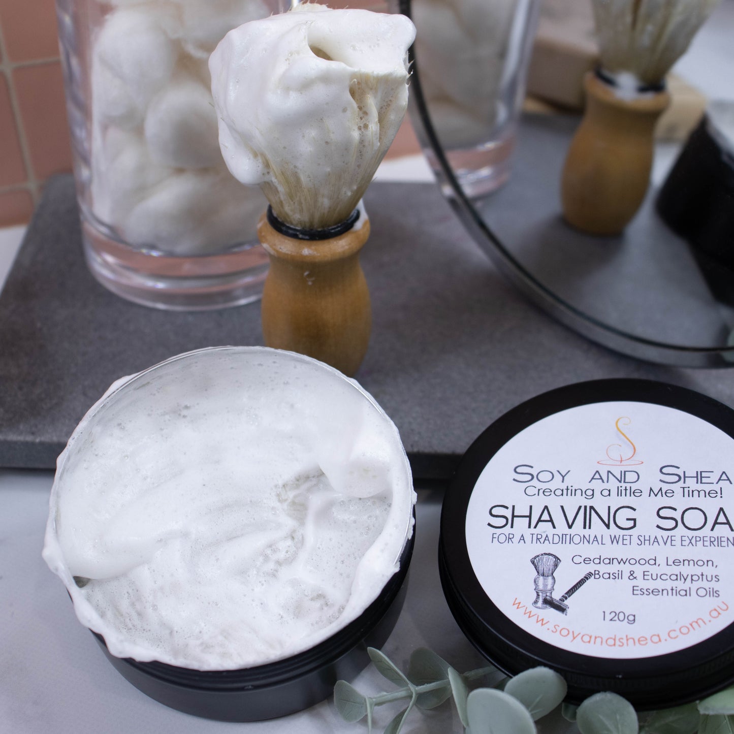 An open black tin of Shaving Soap with a dense lather sits on the side with the lid next to it. Behind s a grey stone tray with a shaving brush standing upright and covered in foam along with a jar of cotton balls and a mirror reflecting the scene.