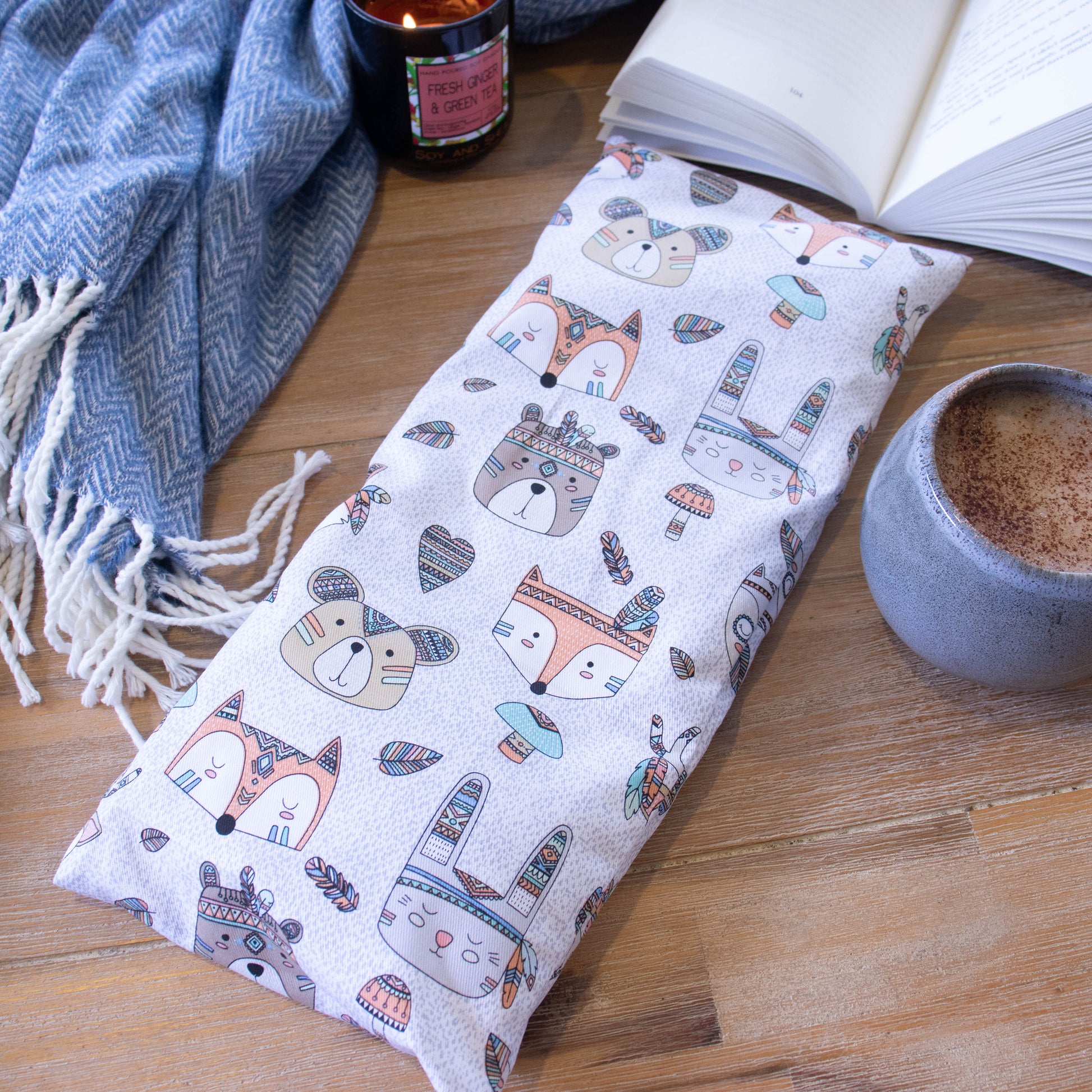 A long rectangular heat pack lies on a wooden surface surrounded by a blue blanket, open book, burning candle and filled coffee cup.  The Heat Pack is white with mottled grey background featuring illustration of animal heads including fox, bear and rabbit.   Each animal has tribal like markings and some wear feather bands around their head.  Addtional feathers, hearts and mushrooms in tribal patters are scattered between the animals.