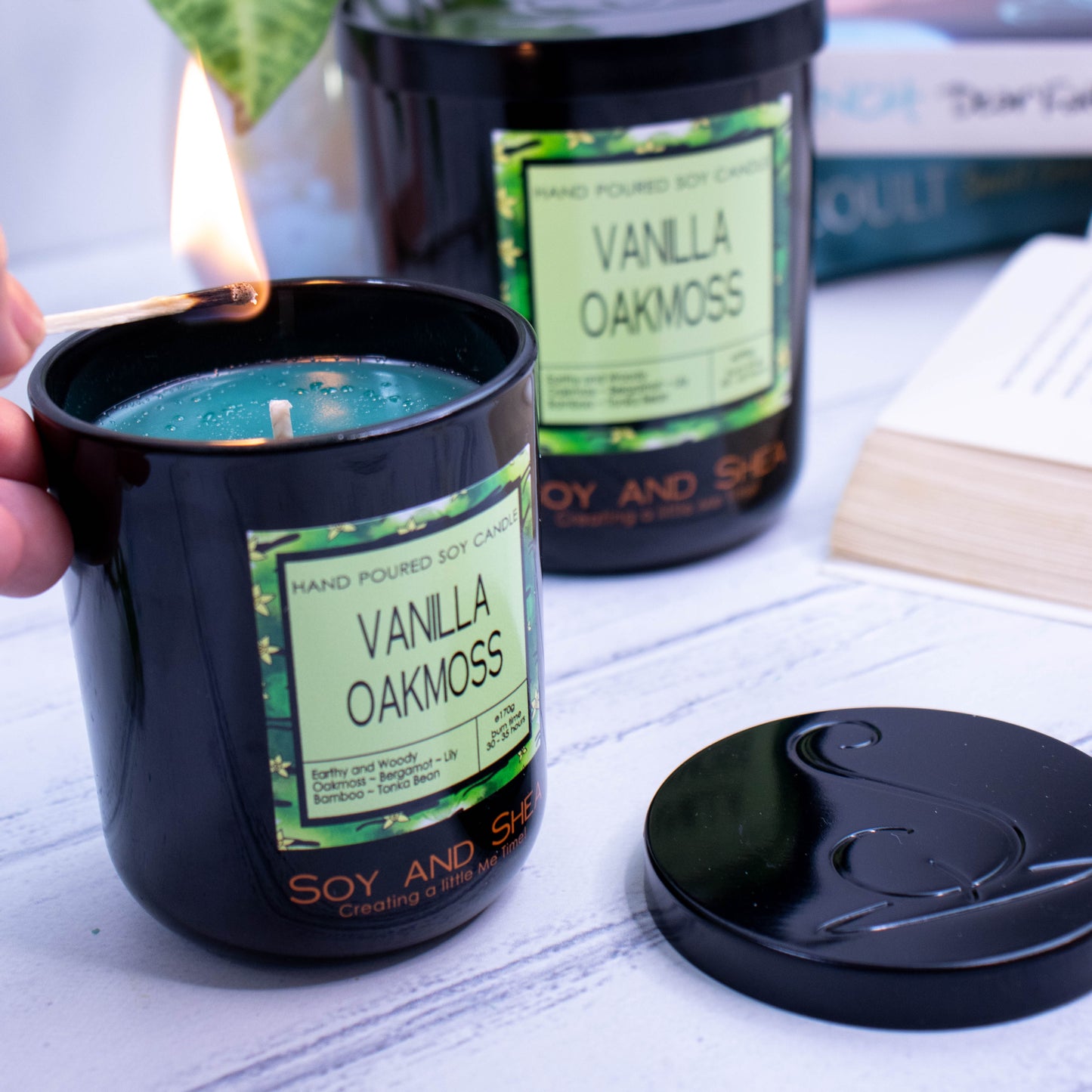Two candles in black jars sit on a wooden bench surrounded by books and plants.  A large candle sits towards the back, while a smaller candle is in the foreground with its lid removed revealling a dark green wax.  A hand reaches in with a lit match.