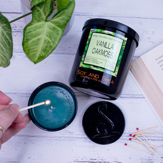 A flat lay photo showing a large black jar lying on its side on a white wooden surface next to a book and plant.  To the bottom of the jar is a smaller candle standing upright and filled with dark green wax.  A hand reaches in with a lit match.