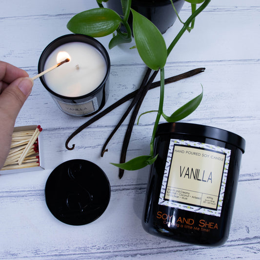 A flat lay photo showing a large black jar lying on its side on a white wooden surface.  To the left top of the jar is a smaller candle standing upright and filled with white wax.  A hand reaches in with a lit match. Surround the candles is a vanilla vine with bright green leaves and three dried vanilla pods