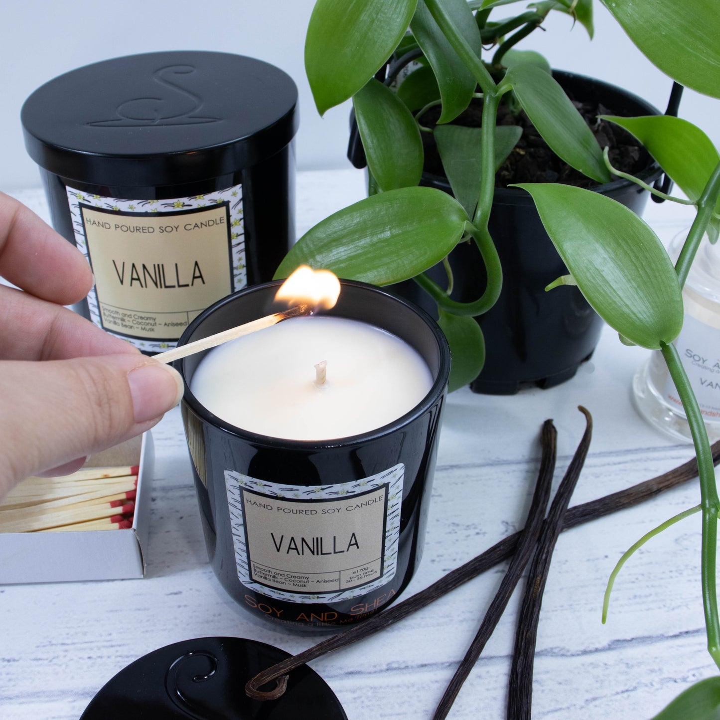 A hand reaches in with a match to light a medium black glass candle filled with a cream wax.  It is surrounded by a vanilla vine and dried vanilla pods.  Behind sits a large candle with a black metal lid with the logomark debossed in its center.