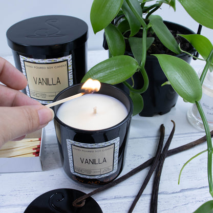 A hand reaches in with a match to light a medium black glass candle filled with a cream wax.  It is surrounded by a vanilla vine and dried vanilla pods.  Behind sits a large candle with a black metal lid with the logomark debossed in its center.
